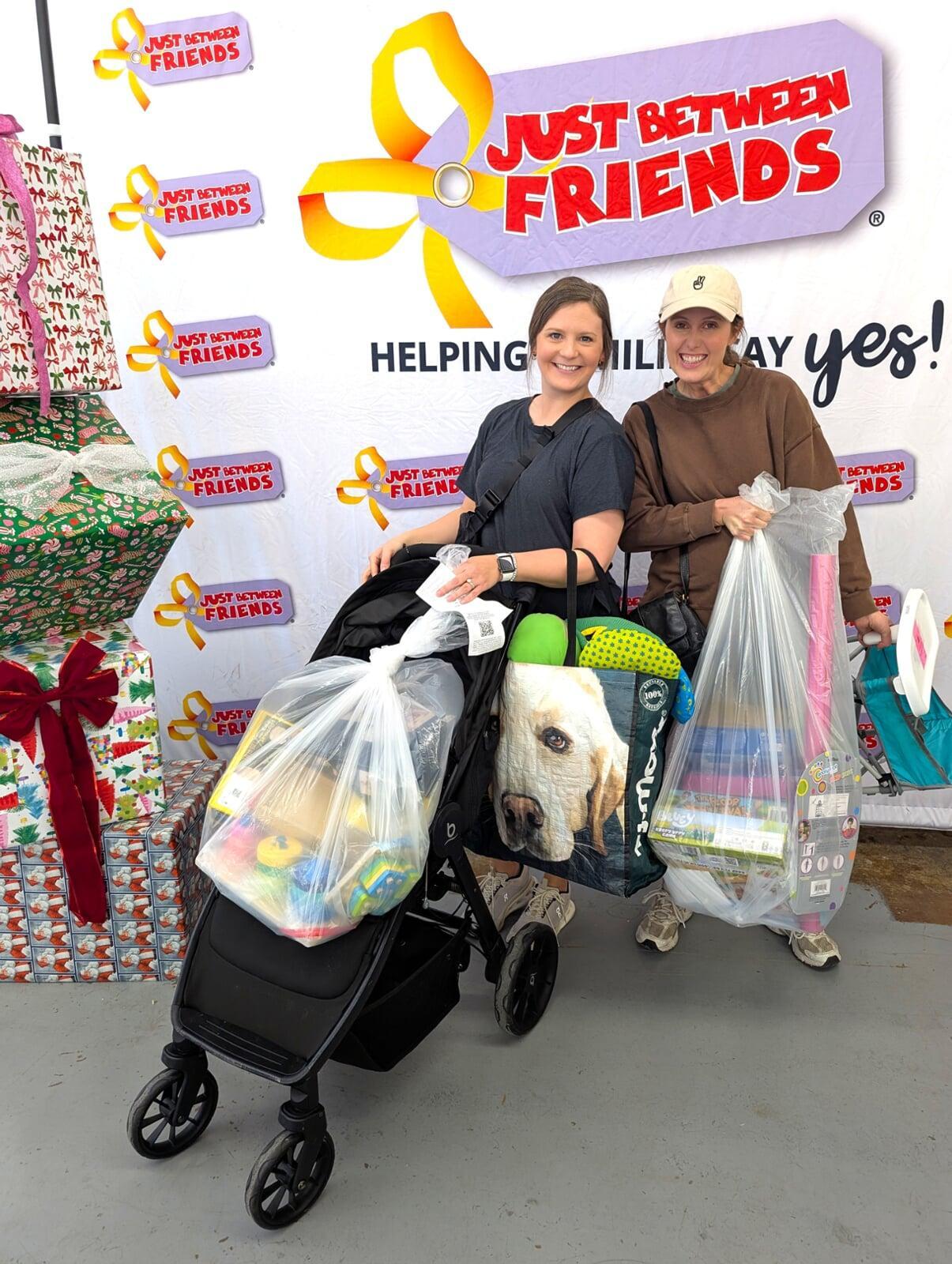 Two women stand in front of a banner that says "Just Between Friends". One is holding a large bag full of toys and games. The other woman is pushing a stroller with two shopping bags. Both women are smiling big.