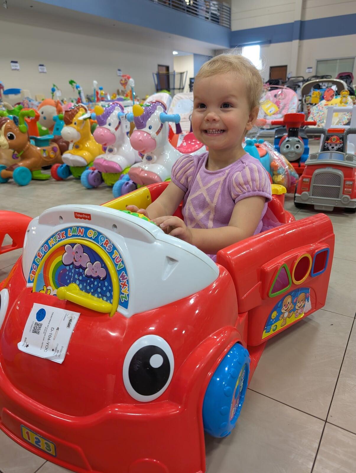A toddler smiles as she sits in a red toy car amid other ride on toys