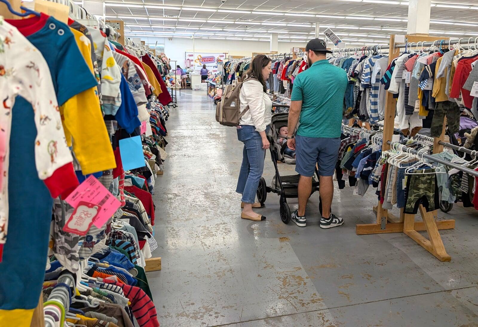 A mother father and baby in stroller stand in a store aisle filled with baby clothes