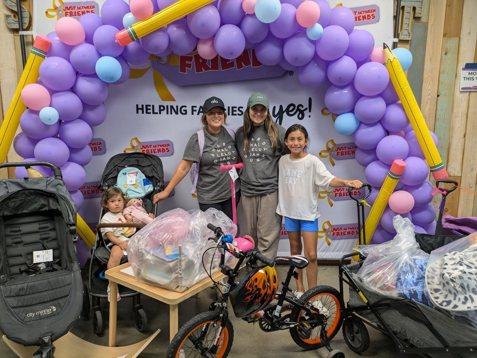 two moms with a school aged child and a toddler in a stroller. They are standing in front of a purple balloon arch with bags of purchases, a cart full of purchases, a new bike and a new stroller.