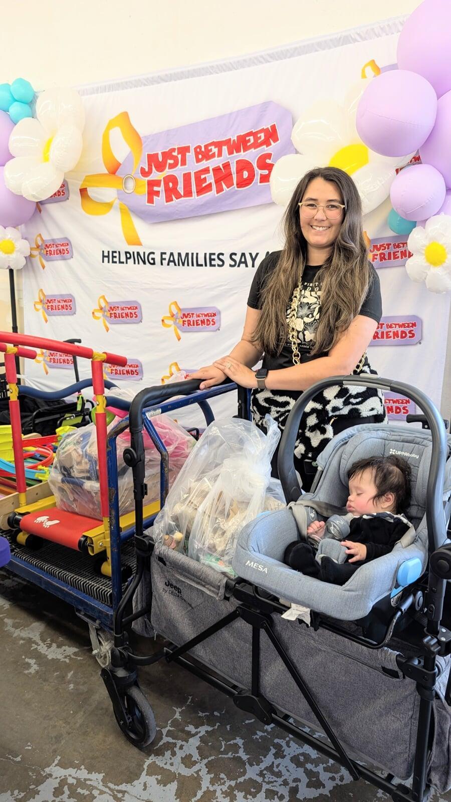 Two women stand in front of a banner that says "Just Between Friends". One is holding a large bag full of toys and games. The other woman is pushing a stroller with two shopping bags. Both women are smiling big.