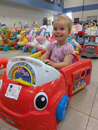 A toddler smiles as she sits in a red toy car amid other ride on toys