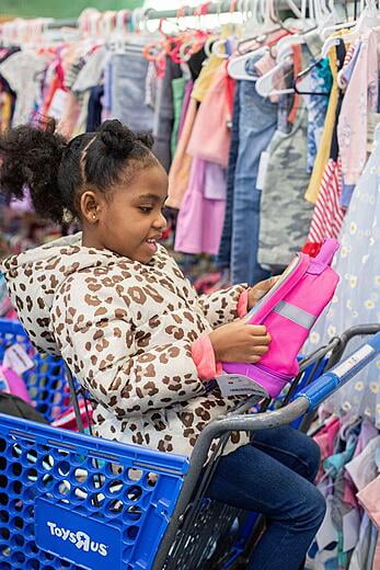 A young girl sits in a blue shopping cart next to a rack of clothing.