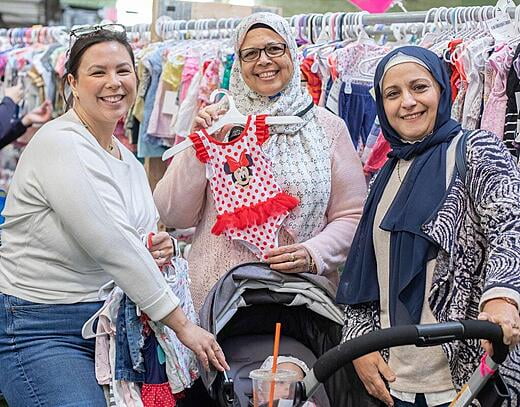 Three ladies stand next to each other in front of a rack of childrens clothing. One woman is holding a bathing suit and they are all smiling.